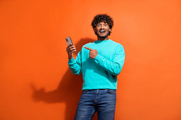 istock Photo of young guy with shock expression wear teal pullover pointing finger his new smartphone features isolated on orange color background 2216135197