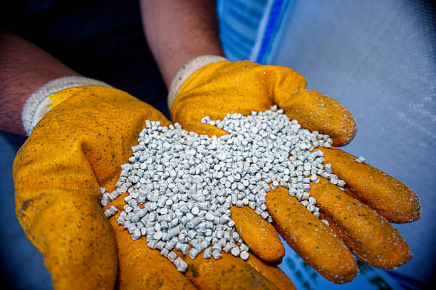 Close-up image of a worker wearing yellow protective gloves holding a handful of white recycled plastic pellets. The granules are used as raw materials in plastic manufacturing and injection molding processes. The image represents sustainability, recycling, and industrial production.