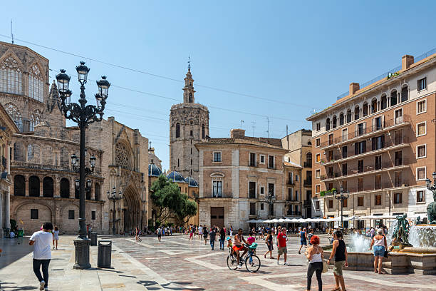 Valencia, Spain - August 12, 2022: Visitors stroll and cycle around Plaza de la Virgen, framed by the Fountain of the Turia and the pink basilica and Gothic cathedral facades.
