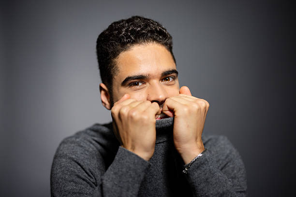 Portrait of young male pulling gray turtleneck sweater over his mouth while looking at the camera. Young man covering part of his face using the neck of his clothing while staring at the camera on gray background.