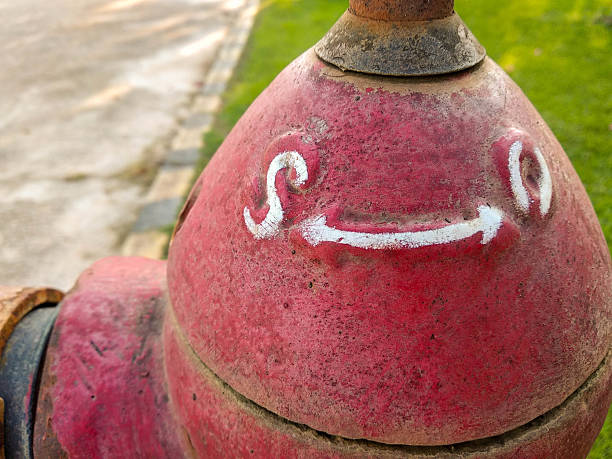 Red fire hydrant top with white painted arrows indicating directions. The surface shows signs of rust and weathering. Positioned near a grassy area and a concrete path, the hydrant is functional and likely used for emergency water access. Positioned at a sidewalk edge, possibly in a public or urban setting.