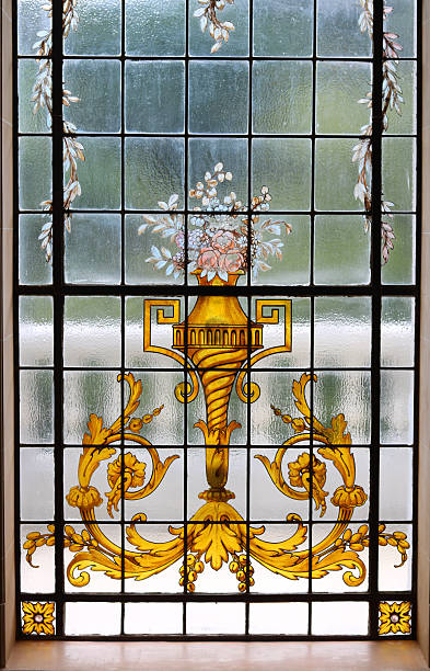 istock High-resolution image of a beautifully crafted stained glass window featuring a classical design with a golden ornamental vase, intricate floral patterns, and pastel-colored flowers. 2216157645