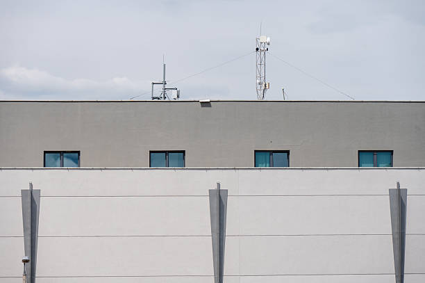 A minimalist industrial building features a clean, geometric facade with small square windows and visible rooftop antennas. The neutral tones and sharp lines convey a modern, functional design often found in commercial or logistical facilities.