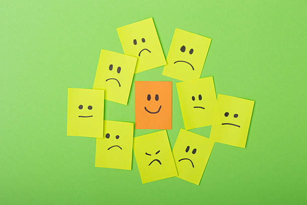 istock A group of yellow sticky notes with sad faces surrounding a single orange note with a happy face on green background. Concept of positivity, individuality, mental health, standing out from the crowd 2216193547