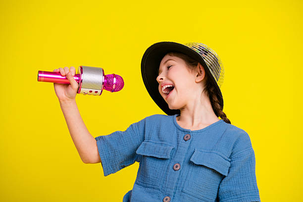 istock Charming young girl enjoying and singing with a pink microphone on a vibrant yellow backdrop, exuding happiness and creativity. 2216211802