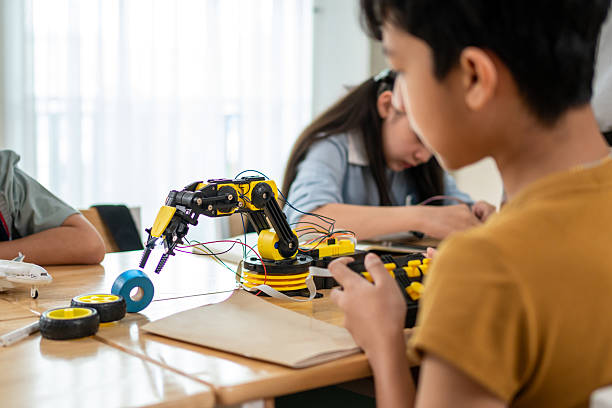 istock Group of students learning in a robotics class at elementary school. Enthusiastic young schoolchildren sitting on table, listening to their teacher explain and teach lesson on robotics in classroom. 2216213133