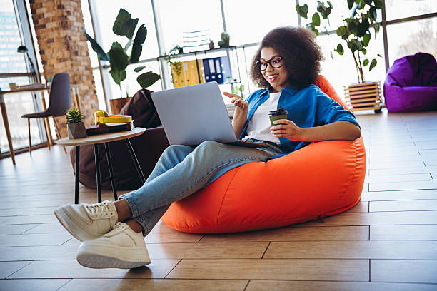 istock Happy young woman enjoying remote work on laptop while sitting in a comfortable bean bag chair in a modern workspace 2216216324