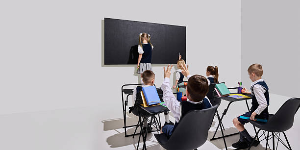 istock Girl writing on blackboard while classmates seated with hands raised and bright books on desks, excited to respond. 2216224878