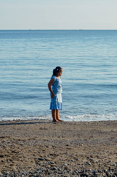A young girl is standing by herself at the seaside after gesturing toward a kite in the sky.