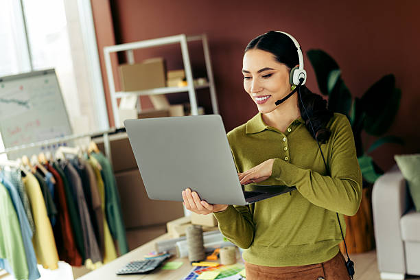 istock Confident young woman wearing a headset working remotely with a laptop in a stylish studio space featuring clothing racks 2216230267