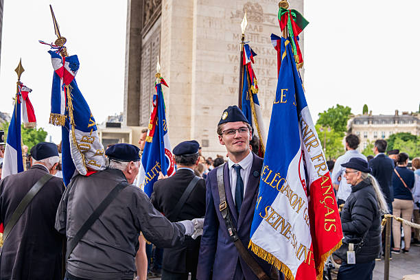 A young man in formal military attire stands among elderly veterans during a commemorative ceremony under the Arc de Triomphe in Paris, France. Holding flags and surrounded by older generations, he symbolizes the transmission of memory and patriotic duty across time.