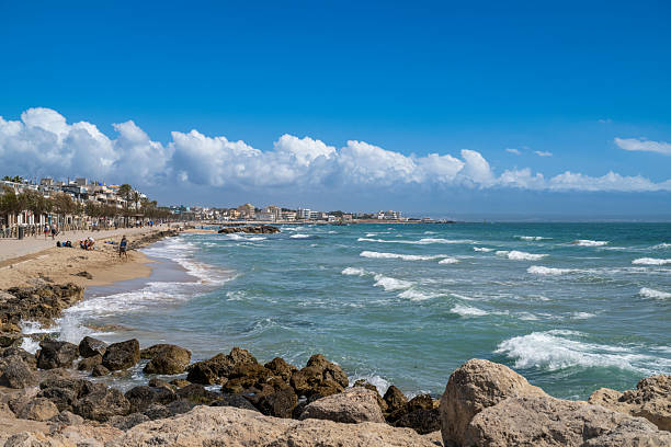 19 April 2025, Palma de Mallorca, Spain. A sunny day illuminates the coastline of Palma de Mallorca, where gentle waves meet a lively beach lined with homes, palm trees, and strolling visitors enjoying the Mediterranean breeze.