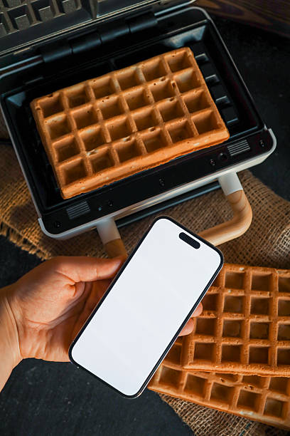 istock Hand holding a smartphone with a blank screen, surrounded by waffle-making ingredients including eggs, flour, milk, sugar, and a waffle iron on a white surface 2216249410