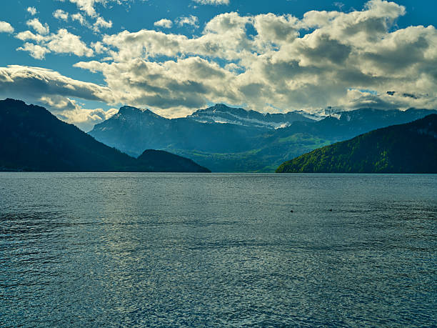 Calm waters of the lake mirror the majestic mountains, surrounded by lush greenery. Fluffy clouds drift across a clear blue sky on a tranquil day.
