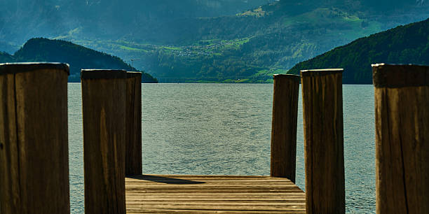 Water flows gently beneath a wooden dock, framed by tall posts. This serene lakeside view showcases a picturesque landscape with rolling green hills and distant mountains in the morning light.