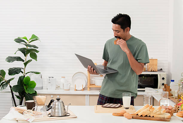 istock Bearded Asian man holding laptop and trying pick up cookies in kitchen while working in morning. Reaching for bread for breakfast on working day. Freelancer working from home and reading news. 2216257321