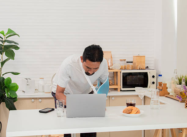 istock Middle-aged man with broken arm and wearing a sling on sick leave working at home in the morning. Sick businessman right arm injury focused looking on laptop computer and using smartphone. 2216257383