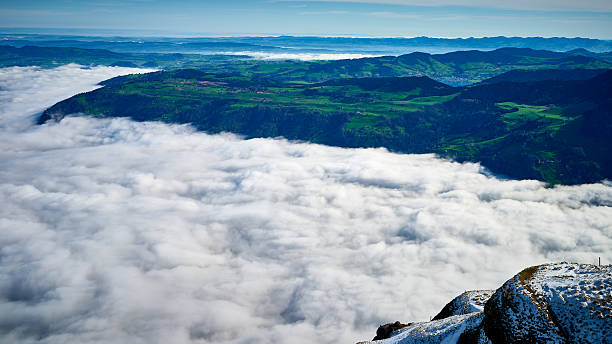 A stunning panorama reveals a mountain peak high above thick clouds, with vibrant green valleys sprawling below. The sun illuminates the landscape, creating a serene atmosphere.