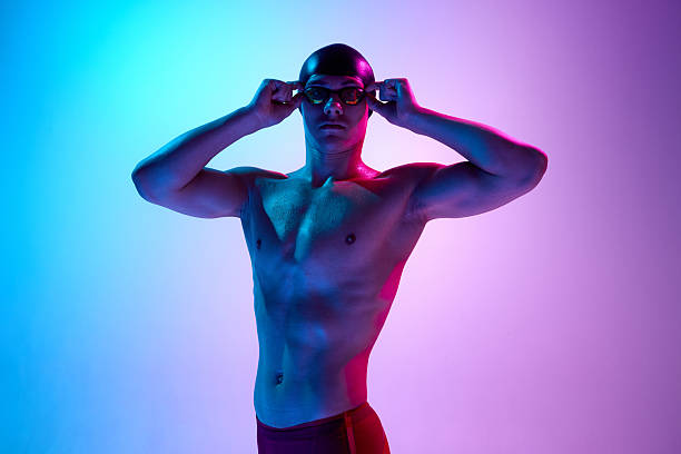 istock Swimmer adjusts goggles with calm expression and intense gaze, body highlighted in vibrant neon light against gradient background. 2216259459