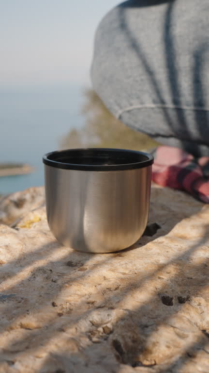 Vertical video. Woman’s Hand with Rings Holding a Metal Cup on a Rocky Surface Overlooking a Coastal Landscape