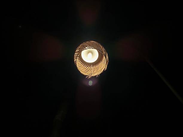 This photo captures a bamboo or rattan-woven lampshade illuminated at night, viewed directly from below. The light bulb at the center emits a warm glow, creating a striking contrast against the surrounding darkness. The pattern of the woven material forms a spiral effect around the light, adding an artistic and cozy touch to the scene. The image evokes a sense of calm and rustic elegance.
