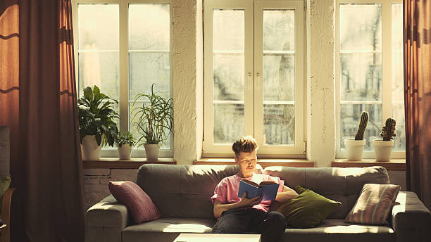 istock Man in pink shirt reads book while relaxing on grey sofa under large bright windows in cozy living room at home 2216263678