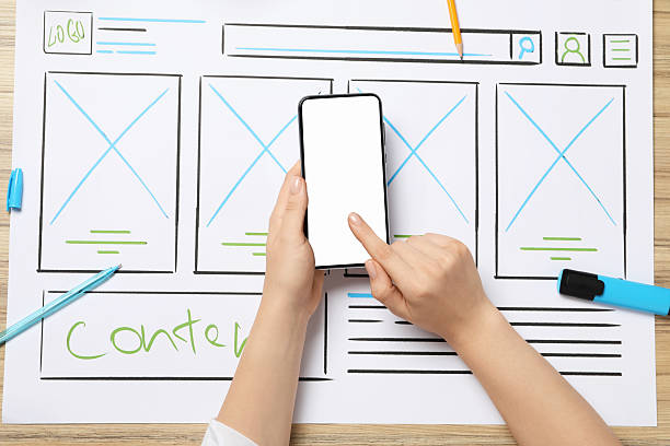 istock Website designer using smartphone at wooden table with wireframe and stationery, top view 2216272503