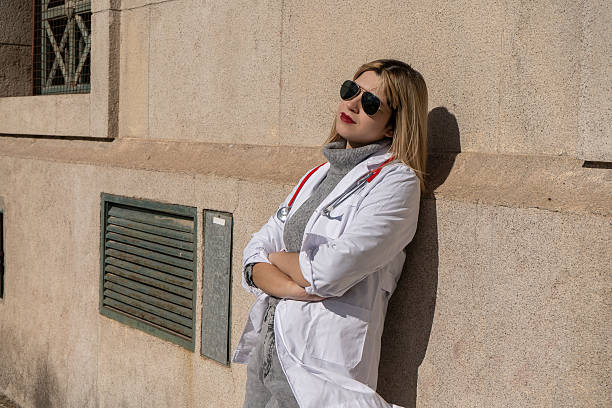 A young female healthcare professional wearing blue medical scrubs is seen walking quickly outside a hospital building, holding her phone and carrying a red backpack over her shoulder. Her focused expression and determined pace suggest urgency  a call to duty or an emergency response. The stethoscope around her neck and ID badge identify her as a nurse, doctor, or emergency medical trainee. This photo captures the fast-paced reality of healthcare  moments when every second matters, and dedication meets responsibility in real-life hospital situations.