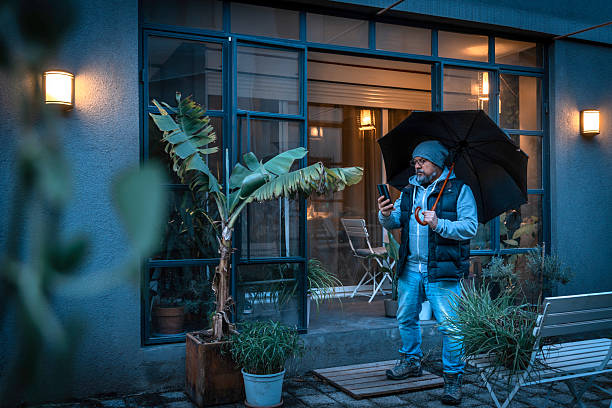 Mature man with umbrella checking his smartphone in front of his house on a rainy evening