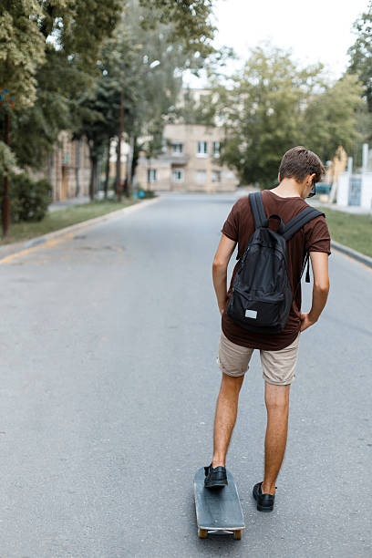 istock Athletic young handsome hipster man in trendy summer clothes with a black backpack rides snowboarding. Fashionable guy enjoys the summer day. Great hobby. View from the back. 2216278330
