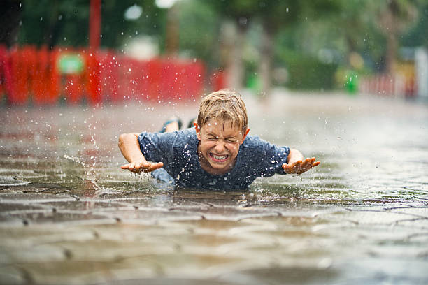 Little boy lying on the wet pavement, fully immersed in the joy of a warm summer rain. With arms spread wide, he appears to be taking a playful splash bath in a puddle, embracing the refreshing droplets falling from the sky. His blue shirt clings slightly to his skin, soaked from the rain, while the surrounding red fencing and greenery add to the serene outdoor setting. \nShot Nikon D850