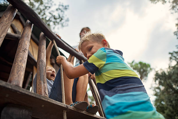 Three children standing on the wooden balcony of a rustic treehouse. The structure is built with wooden beams and railings, blending into its natural surroundings. One boy is climbing the ladder, others are shouting during the play session. The background consists of lush trees and a partly cloudy sky. The kids are enjoying their adventure and exploration in their imaginative outdoor space.Shot with Nikon D850