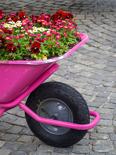 Portrait of an adorable little florist girl in floral dress pushing small wheelbarrow while walking at greenhouse surrounded by flowers and plants.Cheerful small cute girl with wheelbarrow at hothouse