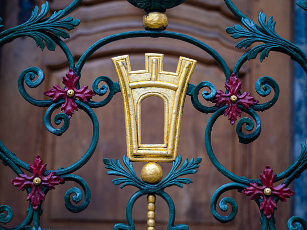 A decorative wrought iron fence featuring intricate floral designs and a central golden emblem resembling a castle tower, set against a wooden background.