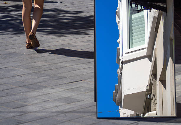 A person walking on a paved sidewalk, with a reflection of a building in a nearby mirror. The scene captures a clear blue sky and urban architecture.