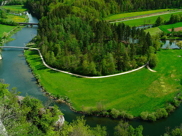 A beautiful natural landscape showcases a river curving past vibrant green fields and dense trees under clear blue skies. Peaceful tranquility reflects in the water.