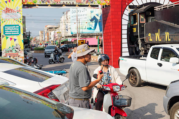 Man with hat and walkie-talkie and sunglasses is standing in street near old thai steam train and locomotoive in front of station in Lampang in  north Thailand. Man is standing between parked cars. A woman is passing by on a motorcycle. In background in fron tof train in main street some traffic is ruling. At buildings in street are big banners for marketing