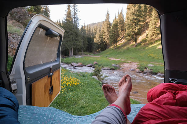 View from inside a camper looking at a mountain summer or spring landscape with spruce forest, blue river and yellow flowers in the morning. Almaty, Kazakhstan. A woman is relaxing with her legs stretched out enjoying the picturesque view