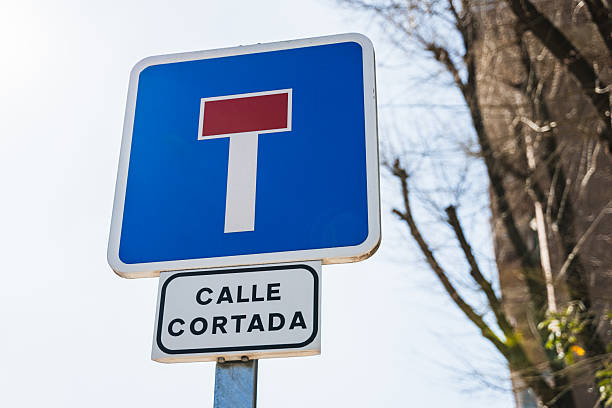 Vertical sign indicating street cut off with text in Spanish. The sign is square with a blue background, a vertical white line and a smaller red line. Below another smaller one with white background and black text. Daylight.