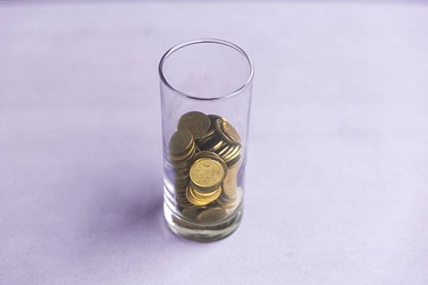 Close-up of a tall clear glass filled with stacked gold coins, set against a soft pastel background. Conceptual image representing savings, finance, investment, and economic growth.