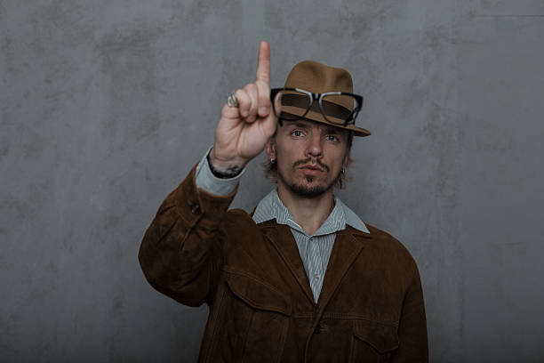 Handsome man hipster raises his index finger up attracting attention. Bearded strong young guy model in a vintage hat in round glasses in a stylish brown jacket posing near a gray wall in the room.