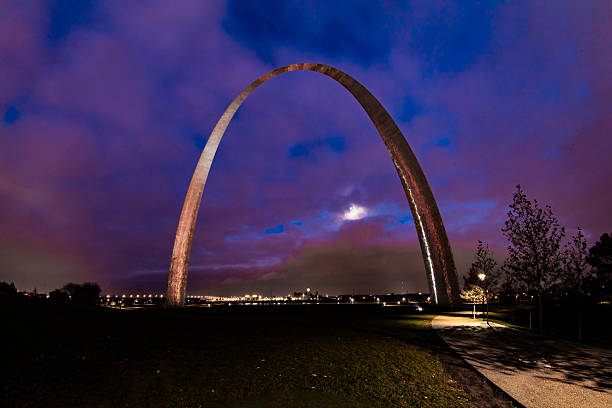 Gateway Arch, early in the evening, shortly after sunset, view is looking east, mostly cloudy sky, moon visible through clouds and low in the sky.  Construction of the arch began in 1963 and was completed in 1965.  Opened to the public in 1967