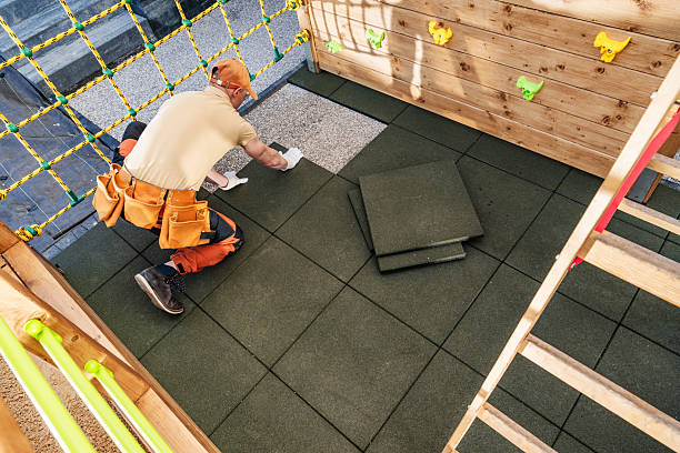 A professional is meticulously laying rubber safety tiles in a playground. The bright environment features climbing structures and colorful wall attachments. The worker is focused on ensuring even placement while using tools to secure the tiles. This care enhances children's play safety and comfort.