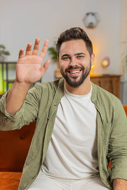 istock Happy young man vlogger smiling friendly at camera and waving hand while sitting on sofa at home 2216351978