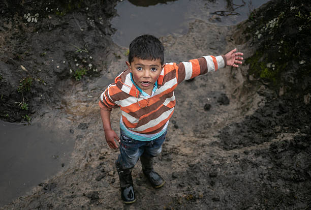 A boy in mud-stained clothes and rubber boots stands on a wet dirt path, raising his arm with a curious and energetic expression. The scene conveys rural life, authentic childhood and connection with nature.