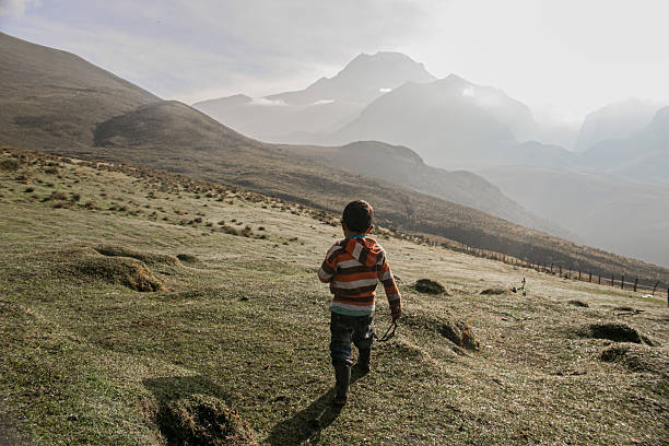 A small boy walks alone over a dew-covered meadow, heading towards the Andean mountains shrouded in morning mist. The scene conveys freedom, exploration and connection with nature in a rural setting.
