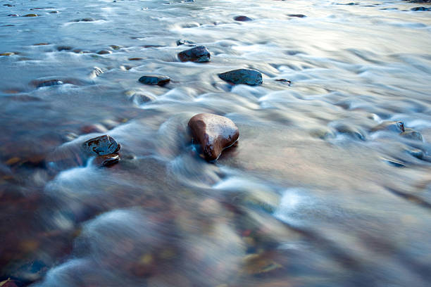 A long-exposure photograph captures the tranquil, flowing water of a clear mountain stream. The movement of the water is rendered as a soft, ethereal blur, creating a sense of peaceful motion. The streambed is filled with a rich variety of pebbles and rocks in shades of brown, grey, and ochre, all visible through the crystal-clear water. The image focuses on the natural textures and serene ambiance of the stream, making it ideal for themes of nature, tranquility, and the beauty of the outdoors.