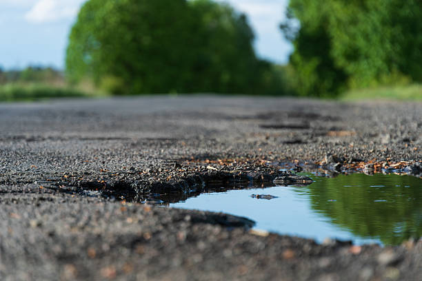 istock Pothole Filled with Water on a Cracked Asphalt Road Surrounded by Green Trees 2216370793