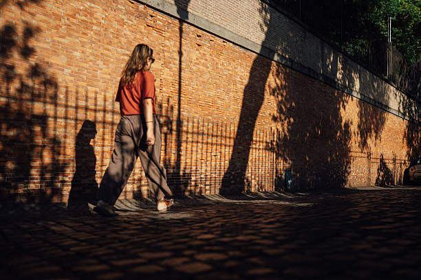 A woman walks along a cobblestone street next to a sunlit brick wall in Rome, with warm afternoon light casting dramatic shadows of trees and railings.