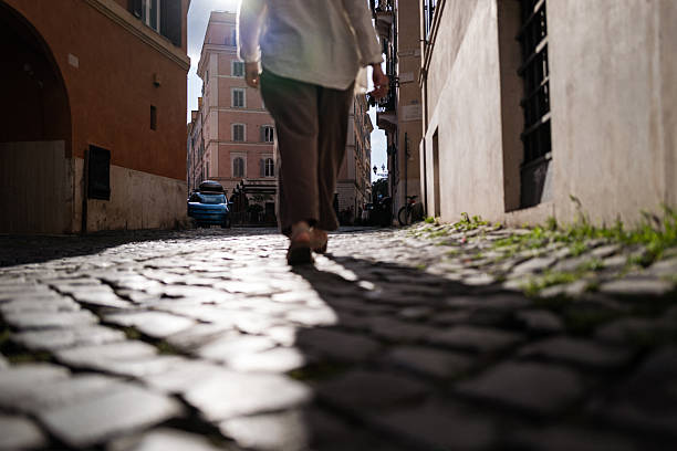 A woman walks along a sunlit cobblestone street in a charming Roman neighborhood, surrounded by pastel buildings with shuttered windows and soft afternoon shadows.